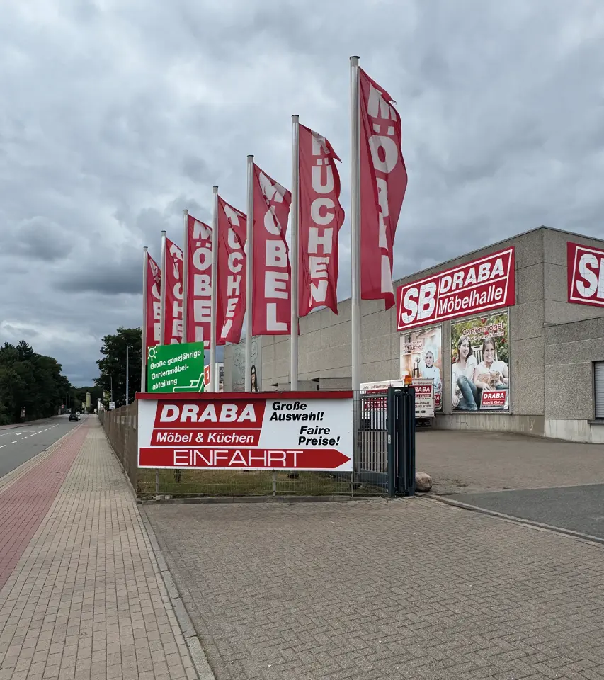 Einfahrt zur SB DRABA Möbelhalle mit roten Fahnen, Schild und Gehweg an der Straße